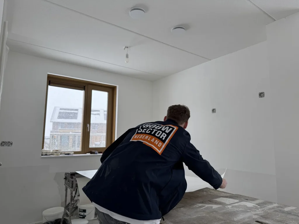 Behanger van Bouwsector Nederland in donkere werkjas rolt een baan vlies uit op de plaktafel, buiten sneeuwt het op de nieuwbouw