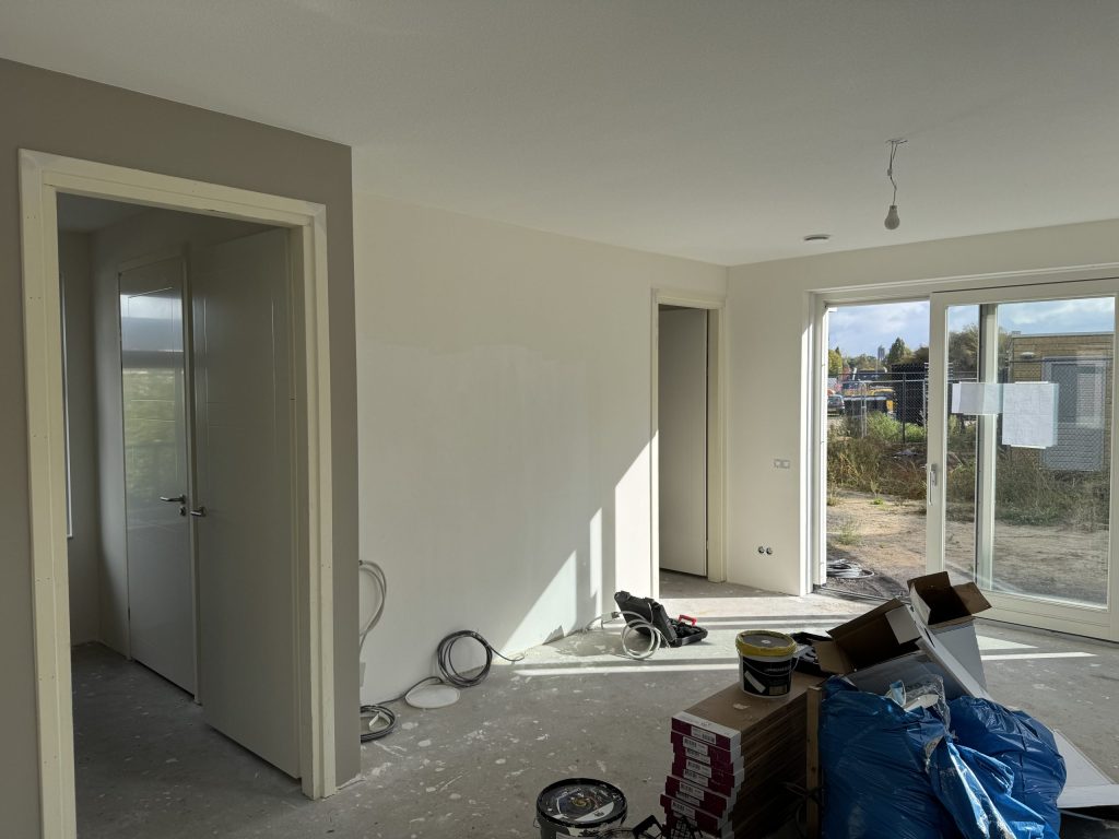 Net opgeleverde woonkamer met kale betonvloer, dozen laminaat en schuifpui naar een tuin die nog moet worden aangelegd
